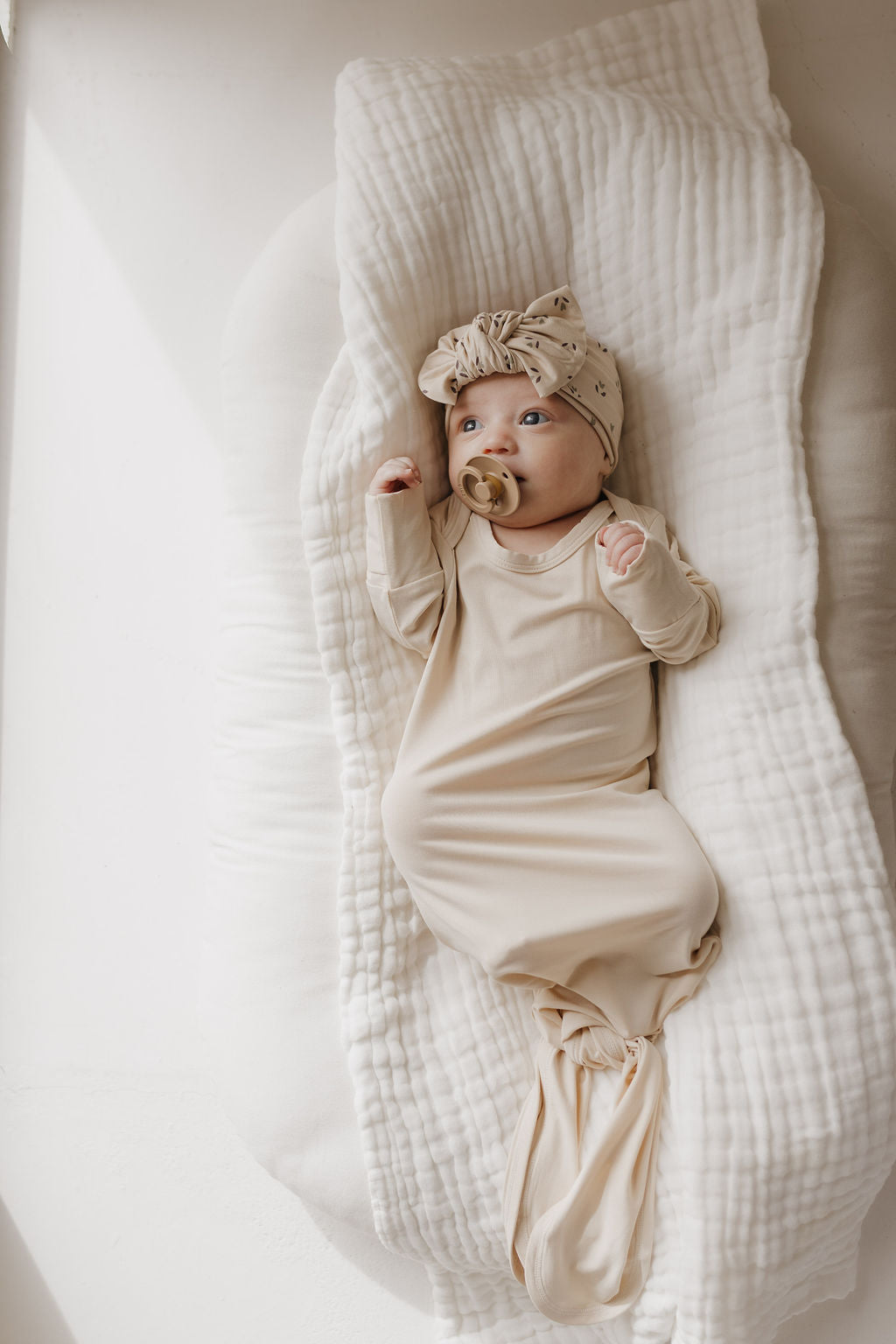 A baby lies on a soft, hypo-allergenic white blanket, dressed in a "Bamboo Knotted Gown | Bone" by forever french baby, with a matching head wrap. A pacifier in their mouth and hands raised slightly, the softly lit scene creates a serene setting perfect for newborn essentials.