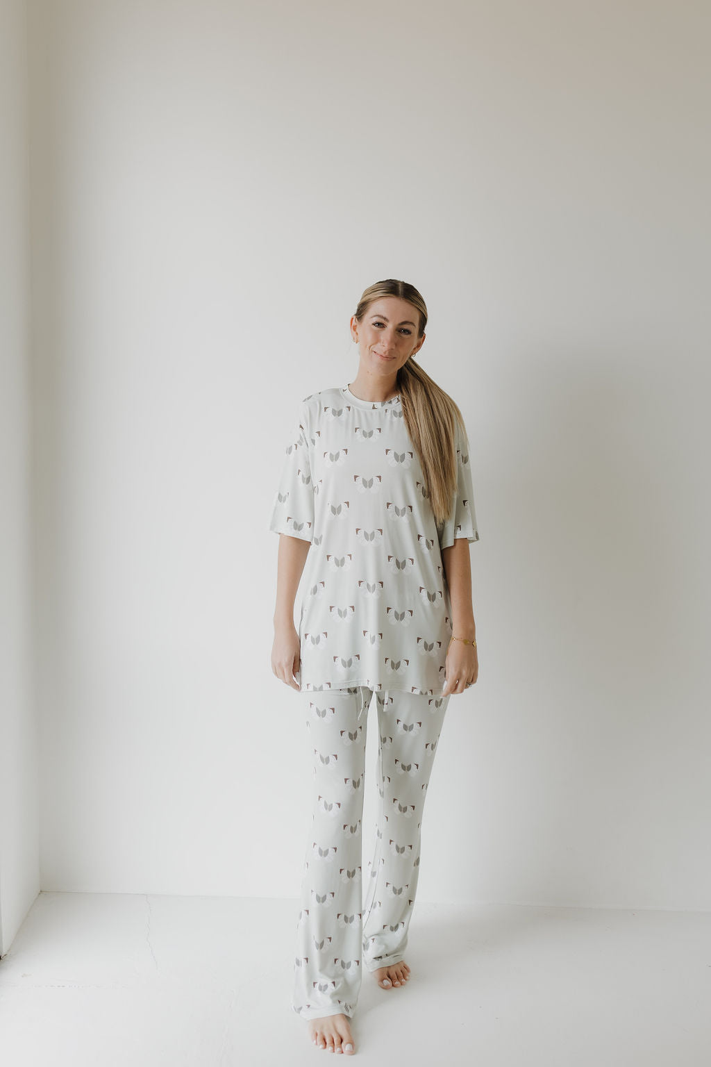 Barefoot on a white background, a person wears an oversized pajama set from forever french baby. The light-colored bamboo pajamas featuring butterfly patterns offer comfort and style, while their hair is styled in a low ponytail.