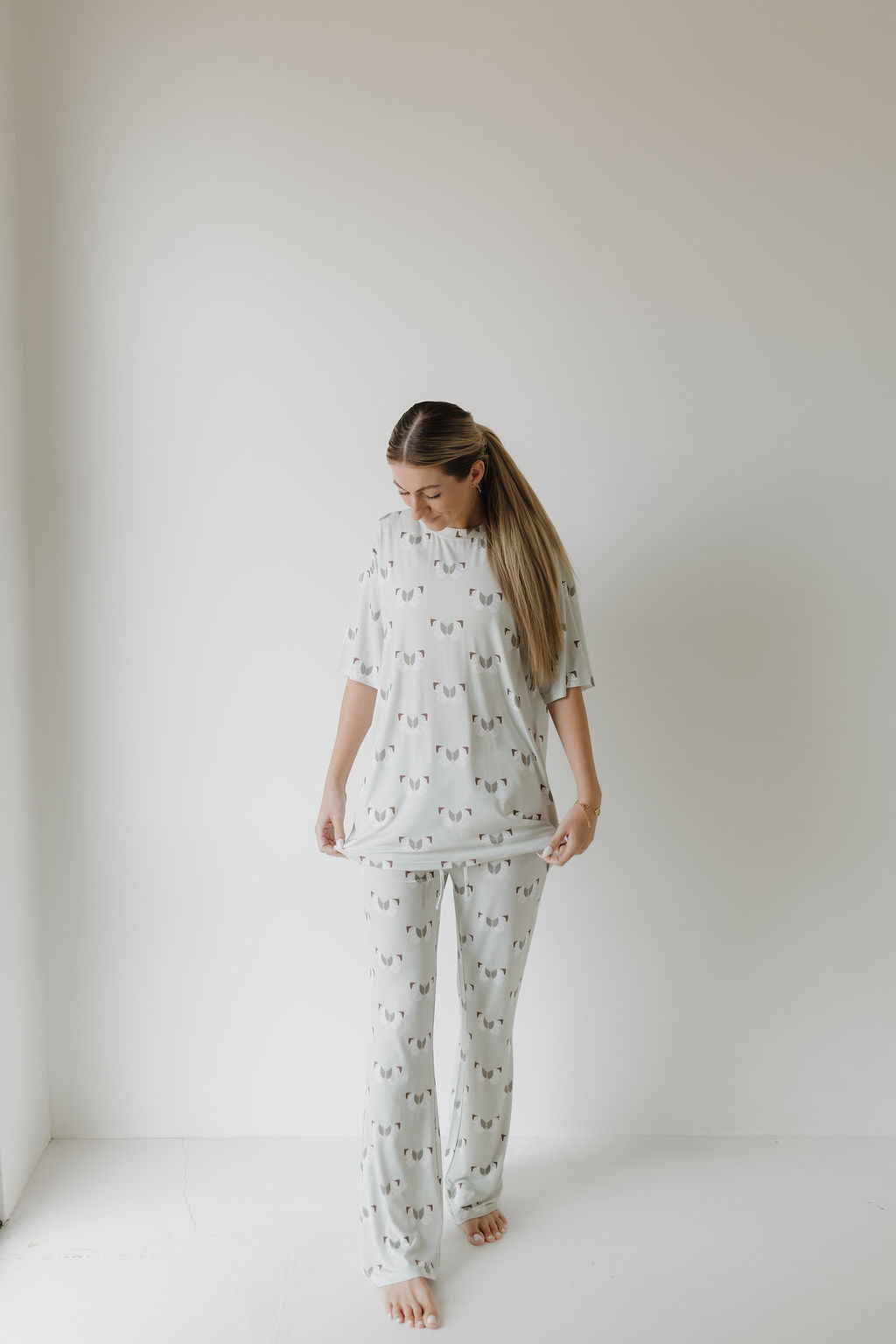 A person with long hair wears the Short Sleeve Women's Bamboo Pajamas | Butterflies from forever french baby. The oversized light gray set, made from hypo-allergenic bamboo, is stylish and comfortable. Standing barefoot in a bright, minimalist room, they stretch the shirt's bottom while looking down.