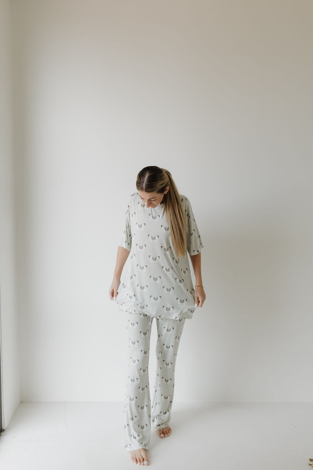 In a minimalist room, a person with long hair stands barefoot, wearing an oversized Short Sleeve Women's Bamboo Pajamas with a subtle "Butterflies" pattern by forever french baby. They look down, holding their top's edge against a light wall, showcasing the hypo-allergenic clothing's soft elegance.