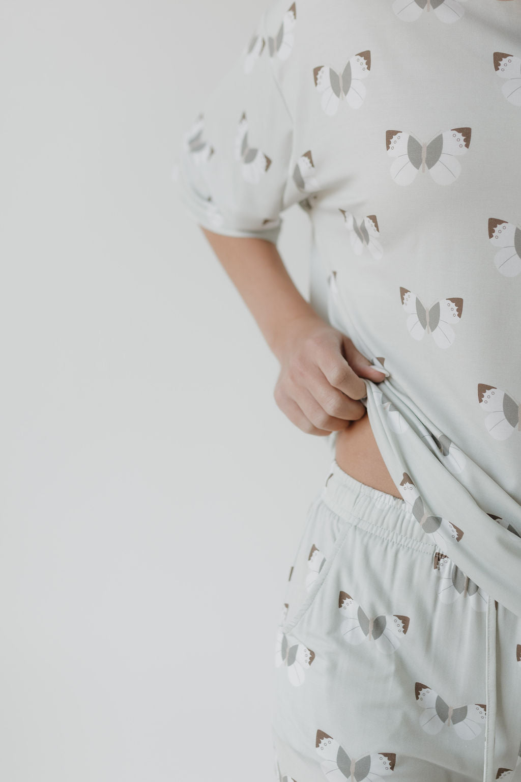 A person is wearing an oversized Short Sleeve Women's Bamboo Pajama set with butterflies from Forever French Baby, subtly lifting the top to reveal their side, against a plain backdrop.