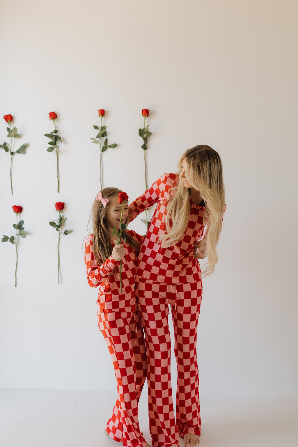 Two people in red and white checkered Youth Flare Bamboo Pajamas by forever french baby stand playfully. One holds a single red rose amid more on the wall. The breathable fabric creates a cheerful, warm vibe against a plain white background.