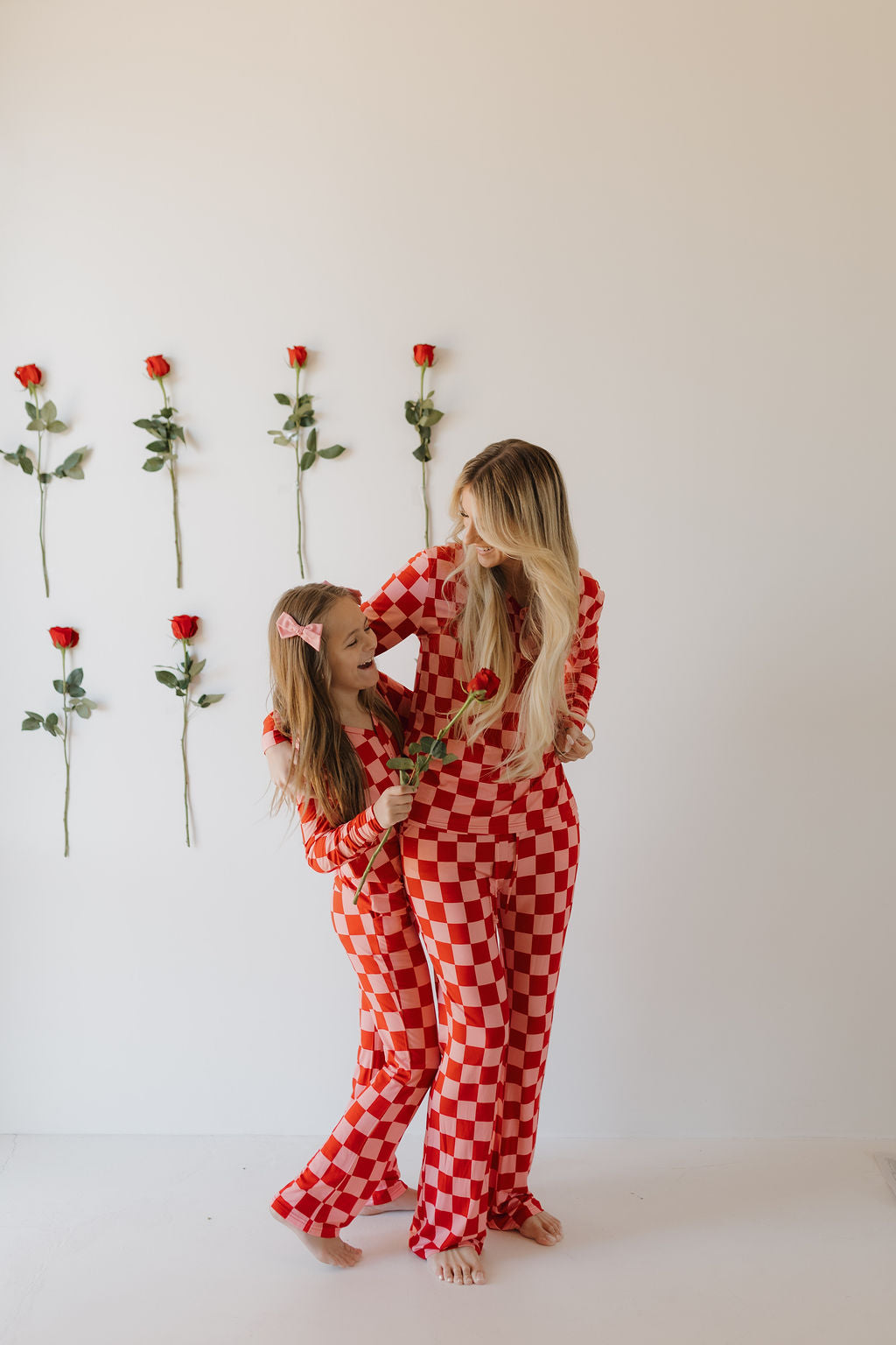 A woman and a girl in matching "Women's Bamboo Pajamas | XOXO" by Forever French Baby, smile and pose together. The girl holds a red rose, and behind them, a white wall is adorned with evenly spaced red roses.