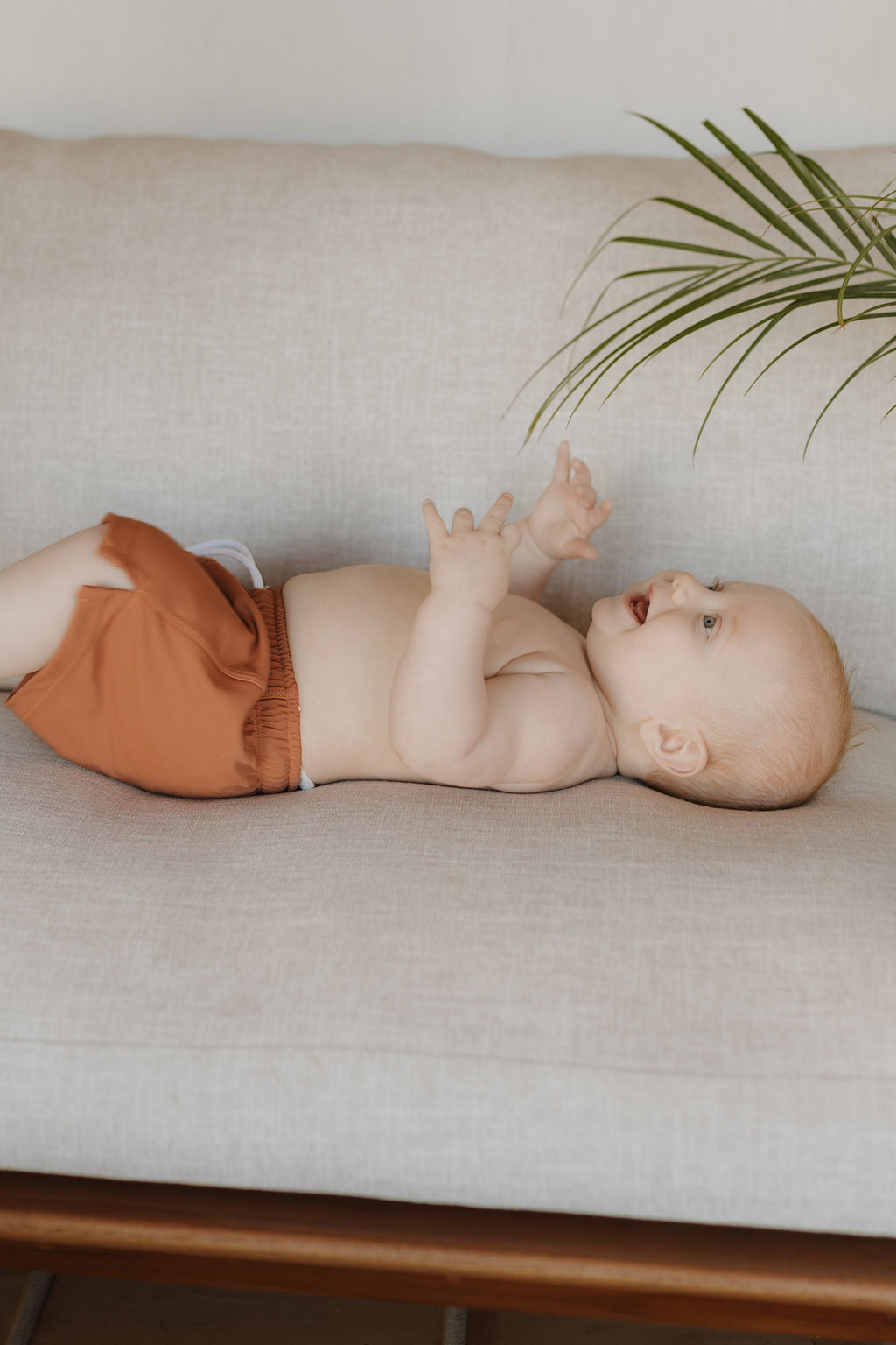 A baby in orange Child Boardshorts from forever french baby's Terra collection lies on a beige couch, reaching upward. A green plant adds a touch of nature to the scene. The happy baby looks engaged, focusing on their hands.