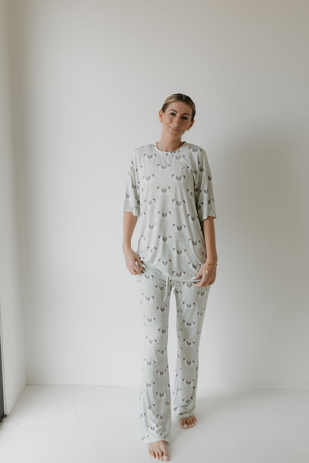 A person stands against a white wall wearing oversized light gray Short Sleeve Women's Bamboo Pajamas | Butterflies by forever french baby, featuring a loose-fit top and pants. Crafted from hypo-allergenic materials, the set complements soft natural lighting as the person remains barefoot.