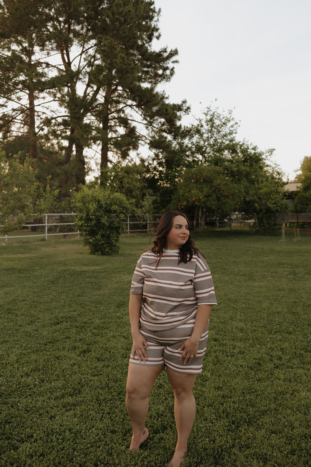 A woman wearing the forever french baby Women's Short Set | Sedona Stripe stands barefoot on green grass in a spacious yard with trees and bushes behind her.