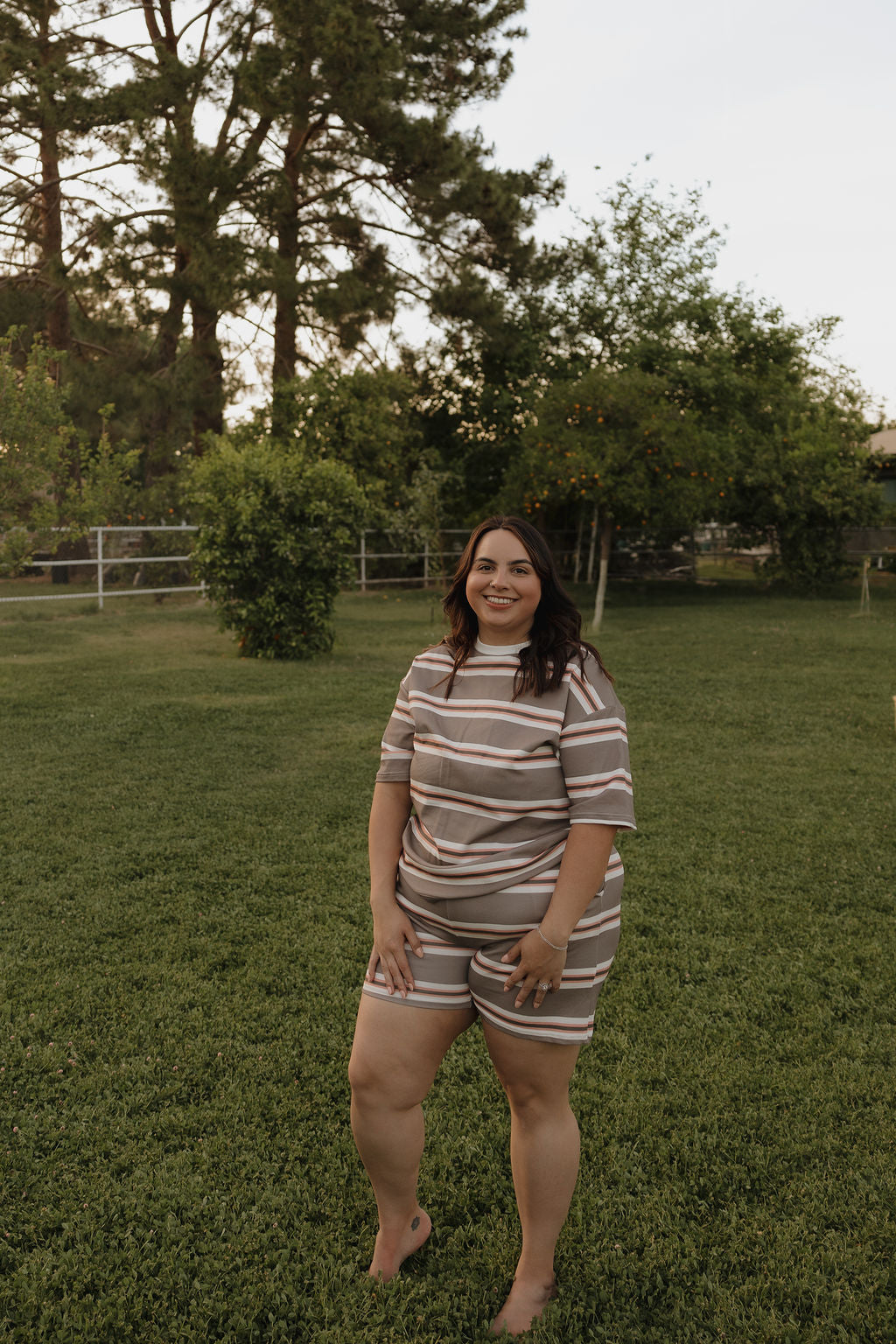 A woman in the forever french baby Women's Short Set | Sedona Stripe stands barefoot on grass in a lush yard, smiling at the camera with trees and bushes behind her.