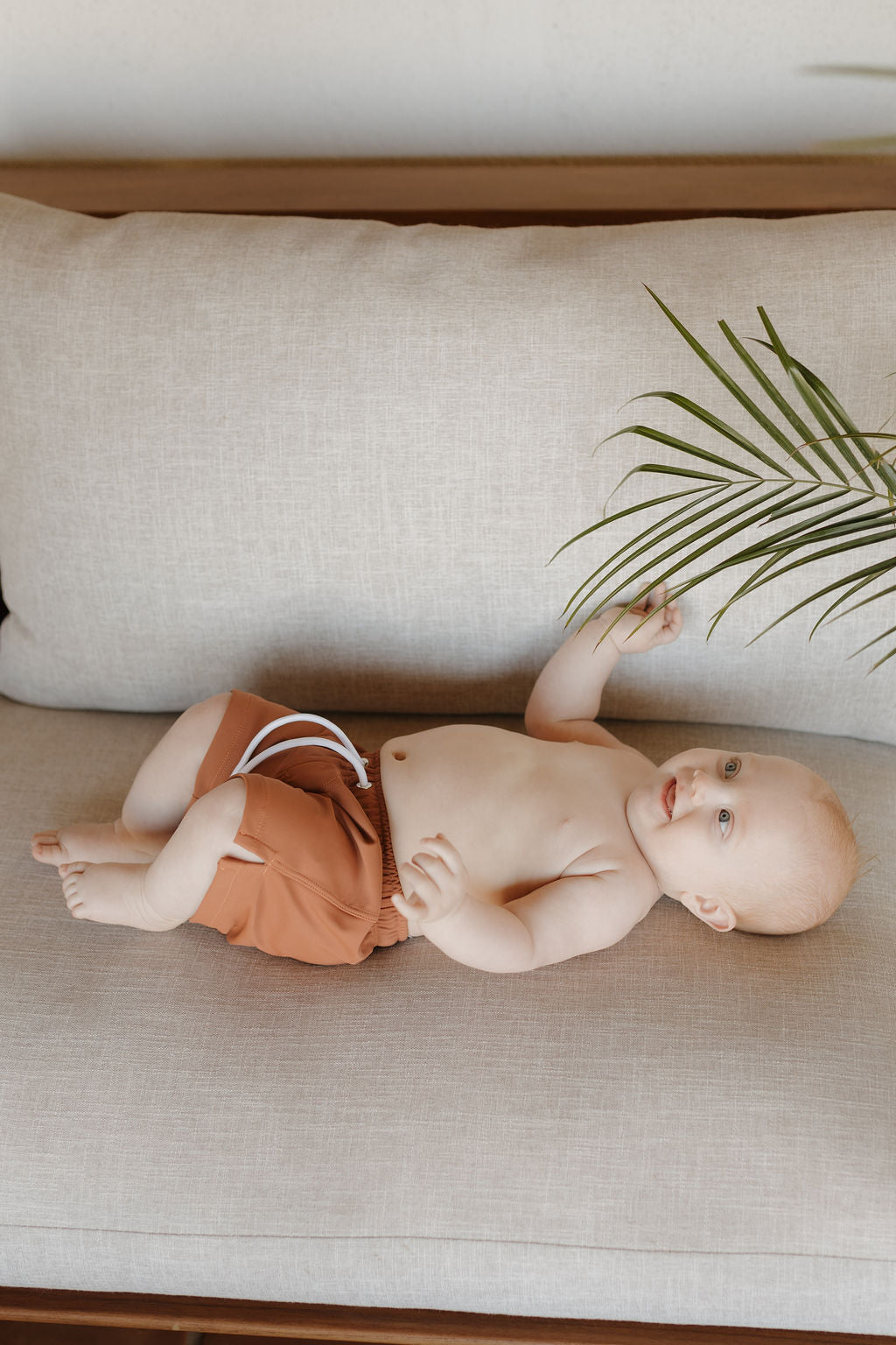 A baby in Terra Child Boardshorts by forever french baby lies on a beige couch, reaching for a green palm leaf. The cheerful expression and natural lighting highlight the warm, earthy ambiance of the Terra collection.