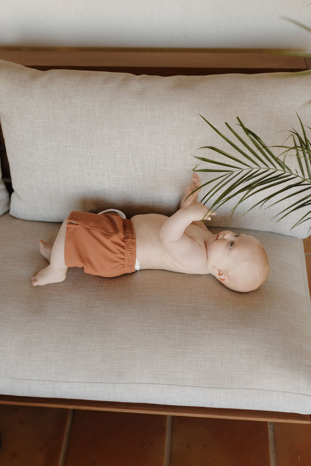 A baby in forever french baby's Child Boardshort | Terra lies on a light gray couch indoors, reaching for a palm frond in a cozy, warm setting.