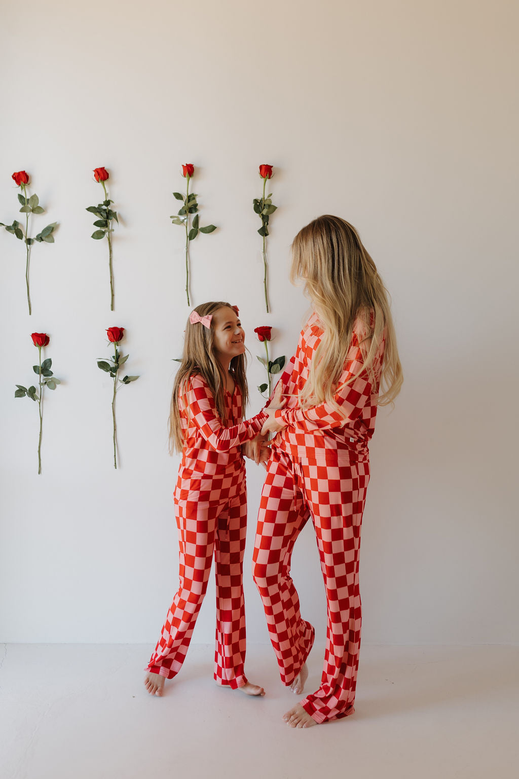 A woman and a young girl stand barefoot, wearing matching forever french baby Youth Flare Bamboo Pajamas in red and white plaid. Smiling, they hold hands against a white wall adorned with evenly spaced vertical rows of red roses.