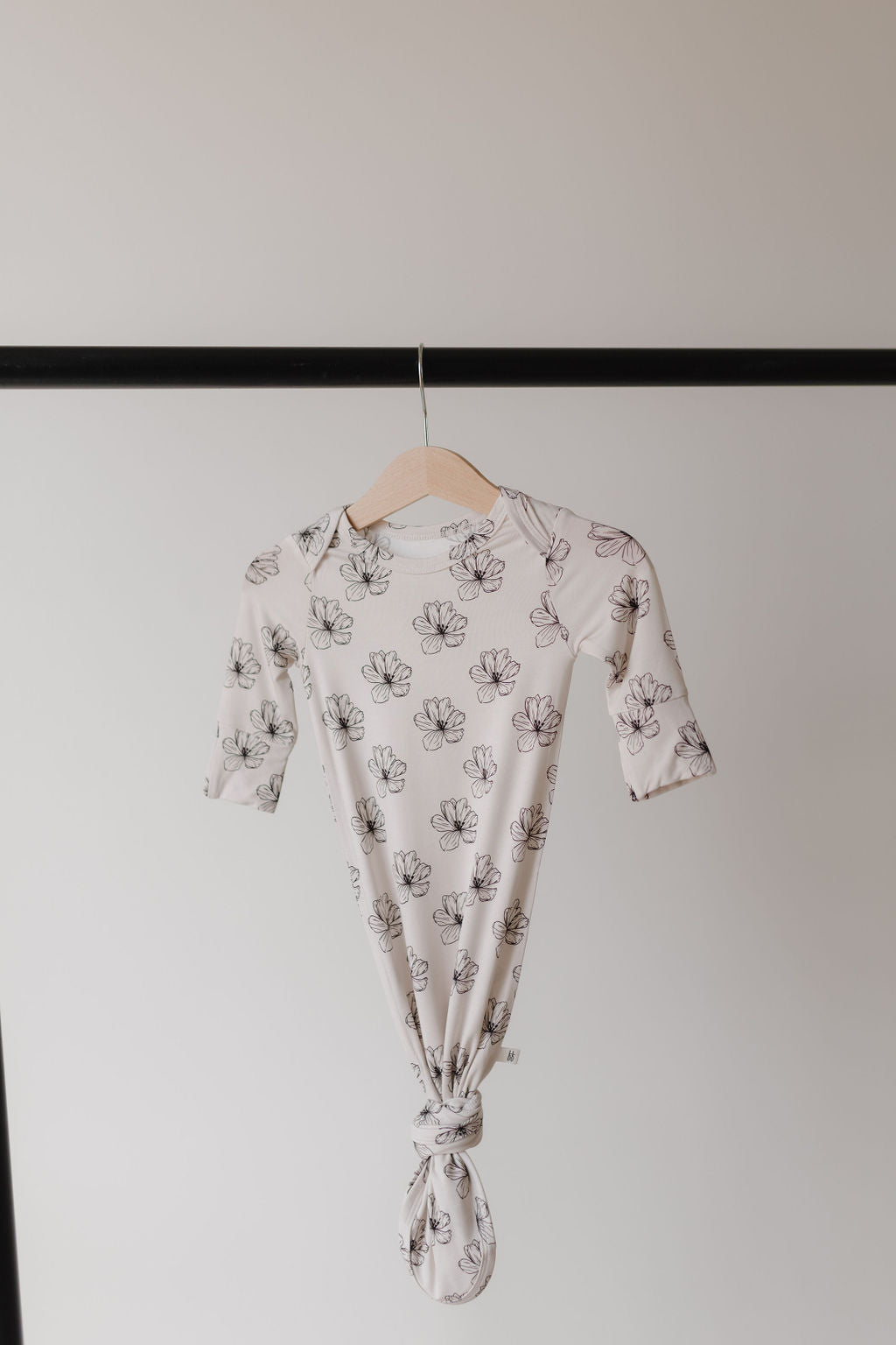 A Bamboo Knotted Gown in Desert Bloom by forever french baby, featuring a black floral pattern on white hypo-allergenic bamboo fabric, hangs on a wooden hanger against a light backdrop. The stylish design ensures comfort for newborns.