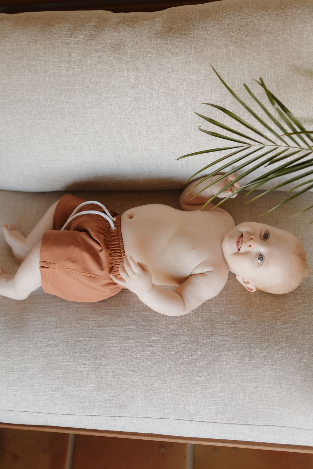 A baby with short, light hair playfully touches a green palm leaf while smiling on a beige couch. The child wears the customizable Child Boardshort from forever french baby's Terra collection. The warm, neutral-toned backdrop enhances the charm of their adorable outfit.