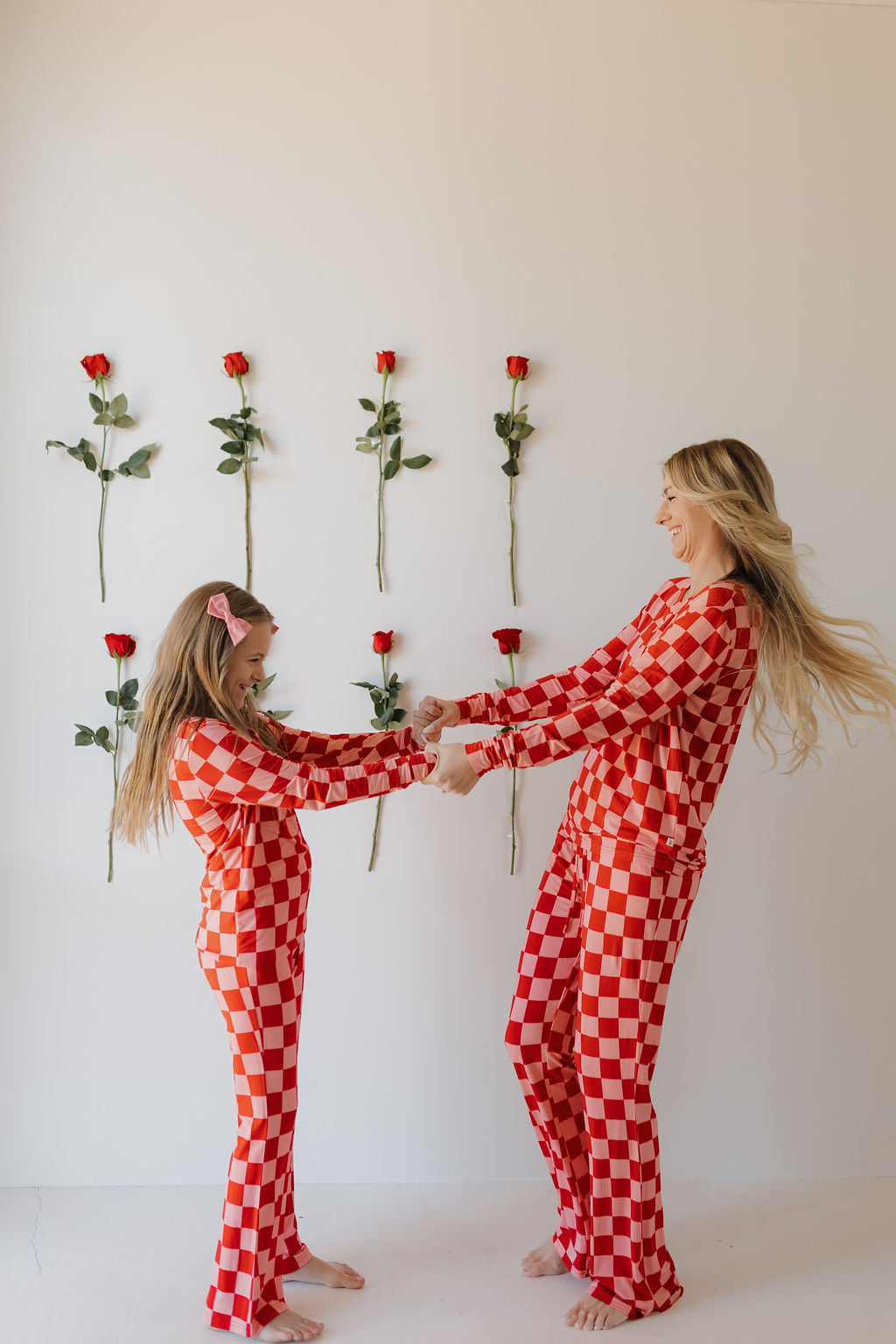 In a minimalistic room, a woman and child wearing forever french baby's Youth Flare Bamboo Pajamas in red and white checkered pattern joyfully hold hands. Six red roses are evenly spaced on the white wall behind them.