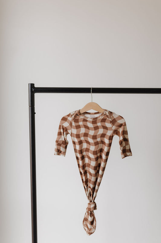 A Bamboo Knotted Gown in Groovy Gingham by forever french baby, featuring brown and white checks, crafted from hypo-allergenic bamboo fabric, hangs on a wooden hanger against a plain white background on a black clothing rack.