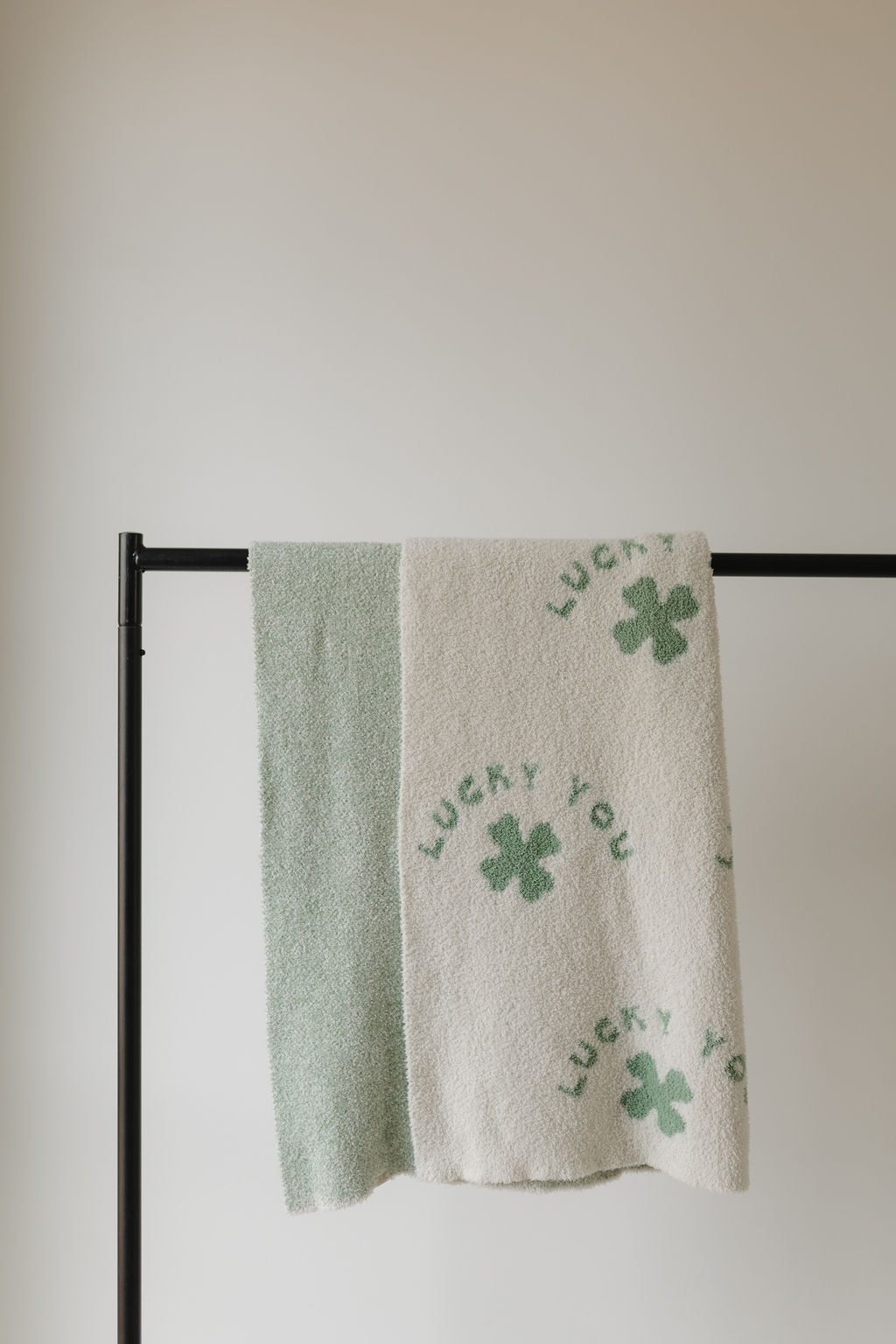 A white Plush Blanket by forever french baby, featuring green shamrocks and "LUCKY YOU" crafted from Oeko-Tex certified materials, hangs on a black metal rod. Beside it is a plain light green towel against a neutral, light wall. (Pre-Order Ships 2/24).