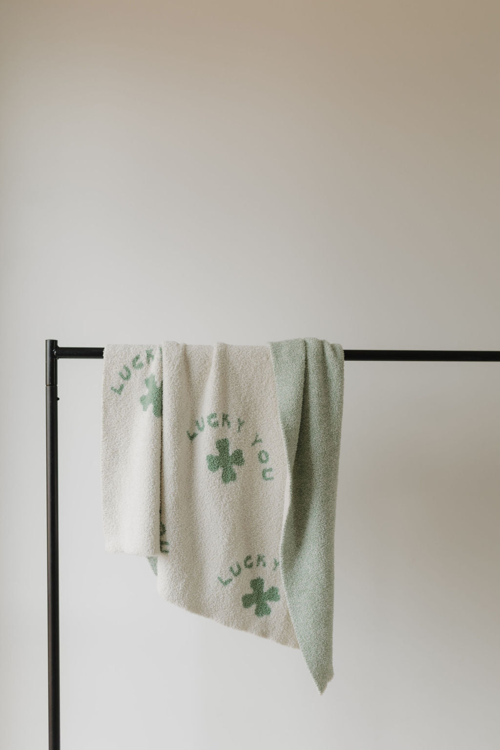 Two towels hang on a black metal rod: the left is beige with "Lucky You" and shamrocks, made from plush microfiber feather yarn by forever french baby, and the right towel is solid light green. An off-white wall in the background creates a tranquil vibe.