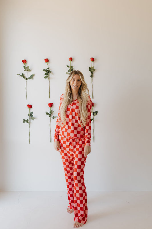 A person in the forever french baby Women's Bamboo Pajamas | XOXO, featuring a red and white checkered design, smiles barefoot against a white wall adorned with six single red roses, highlighting the breathable fabric.