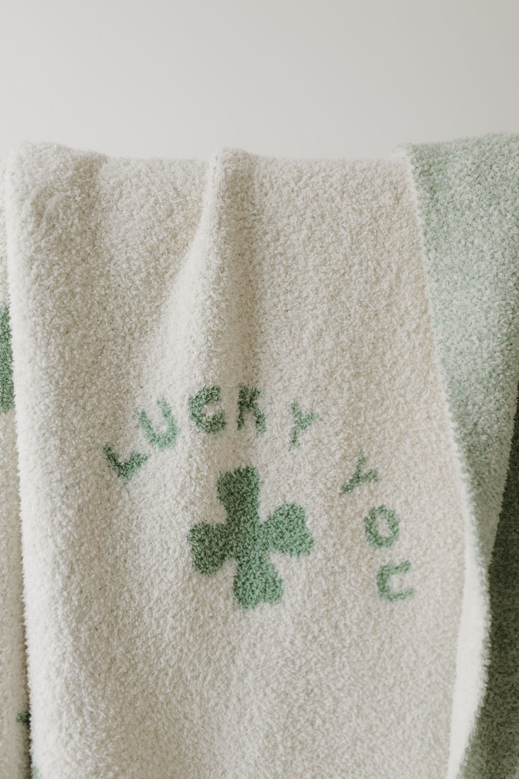 The Plush Blanket | Lucky You by Forever French Baby, crafted from Oeko-Tex certified microfiber feather yarn, features "LUCKY YOU" text above a four-leaf clover. A subtle green stripe pattern adds elegance. Pre-order now to ship by 2/24.