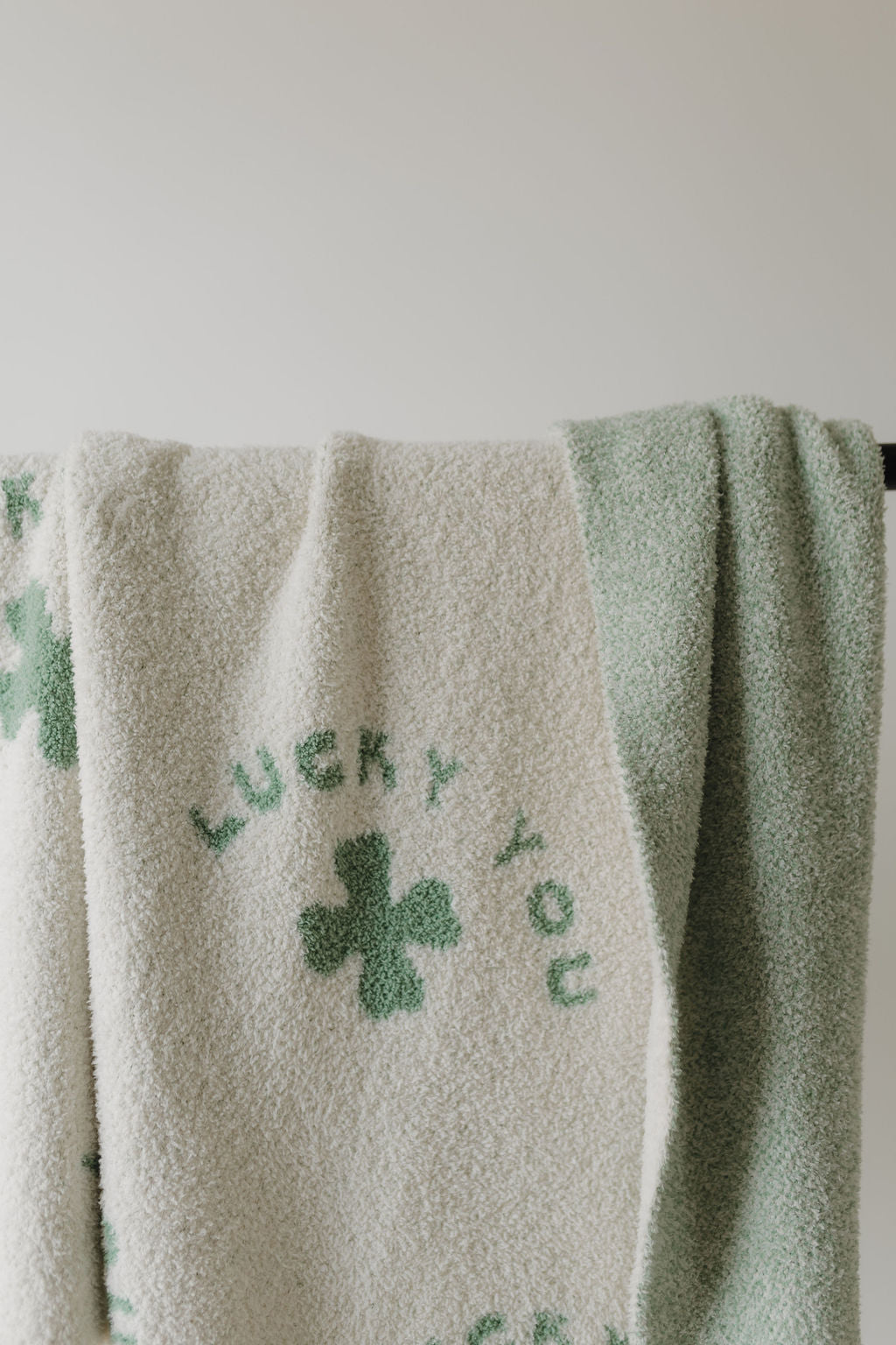 A Forever French Baby pre-order plush blanket, shipping 2/24, hangs elegantly. Crafted from microfiber feather yarn, it features a green four-leaf clover and "LUCKY YOU" on white with mint green patterns and Oeko-Tex certification for safety and style.