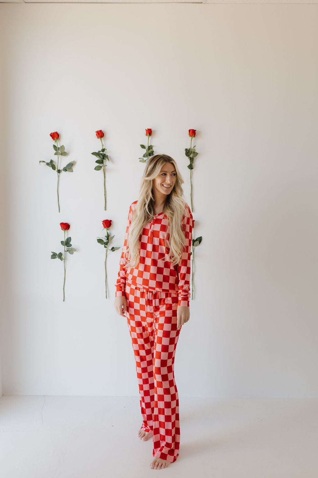 Barefoot by a white wall, a person with long wavy hair wears forever french baby's Women's Bamboo Pajamas | XOXO in red and white checkered pattern. Behind them, simple red roses are aligned vertically.