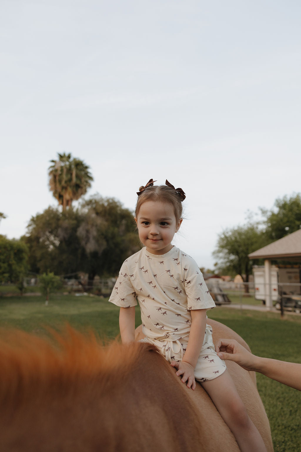 A young girl in the forever french baby Girl's Bamboo Short Set | Wild Horses sits on a brown horse outdoors, supported by an adult’s hand, with trees and a building in the background.