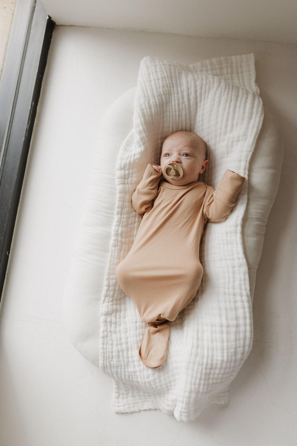 A baby rests in a light-textured cocoon, dressed in forever french baby's soft, hypoallergenic Bamboo Knotted Gown in Tawny. Holding a pacifier and surrounded by soft window light, the infant embodies minimalistic newborn essentials.