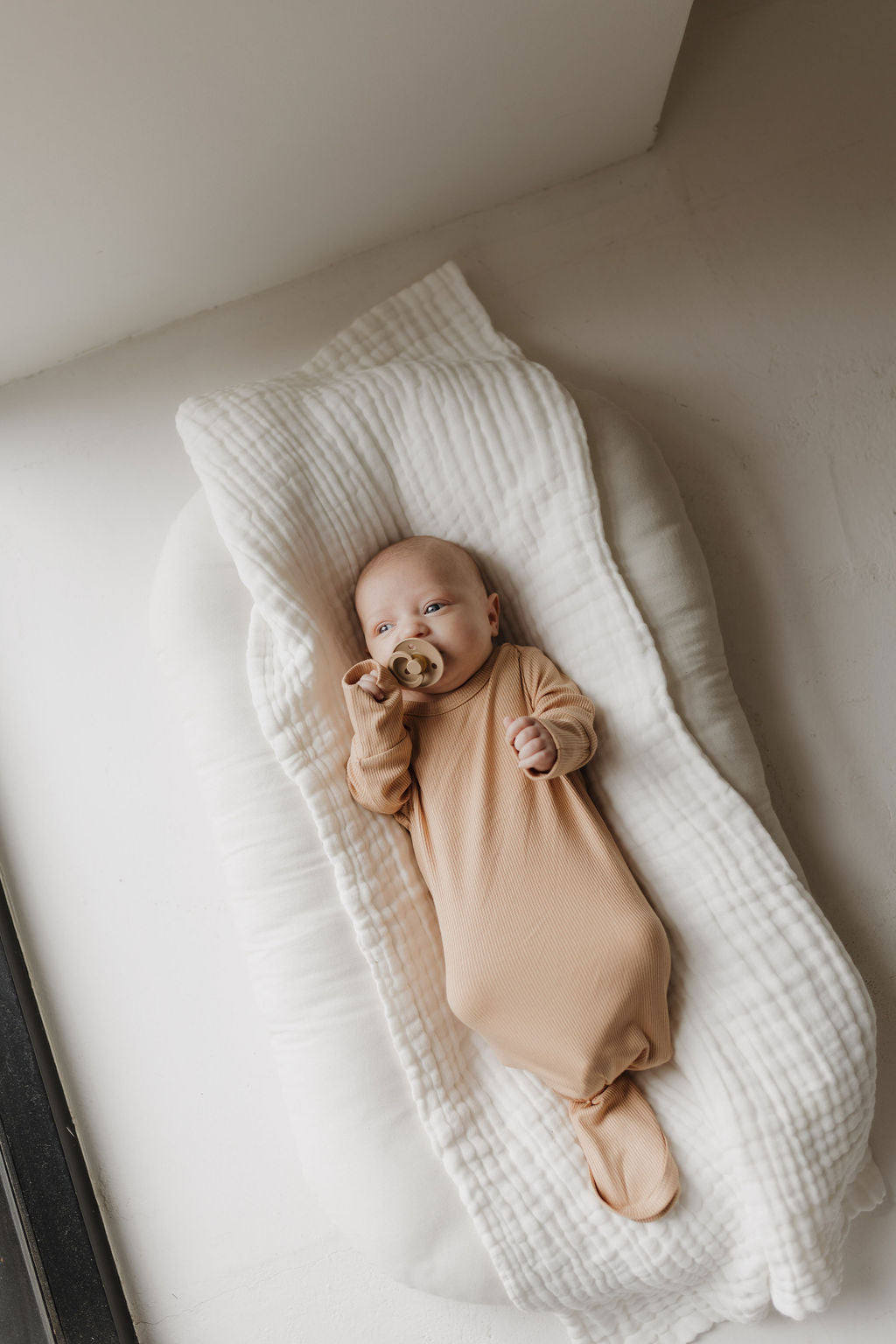 A newborn in a forever french baby Bamboo Knotted Gown | Tawny lies on a cushioned, white blanket by a window. Holding a pacifier, the baby basks in natural light, which softly illuminates the serene scene.