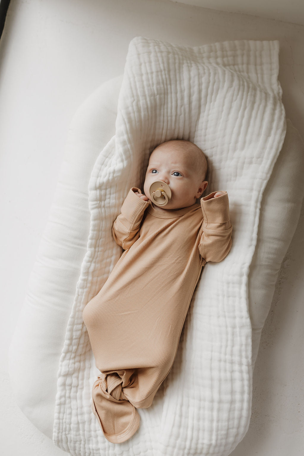 A baby with a pacifier lies on a white cushion, wrapped in the Bamboo Knotted Gown | Tawny by forever french baby. Made from hypo-allergenic bamboo fabric, the gown enhances the serene ambiance as the curious baby looks up against a softly lit backdrop.