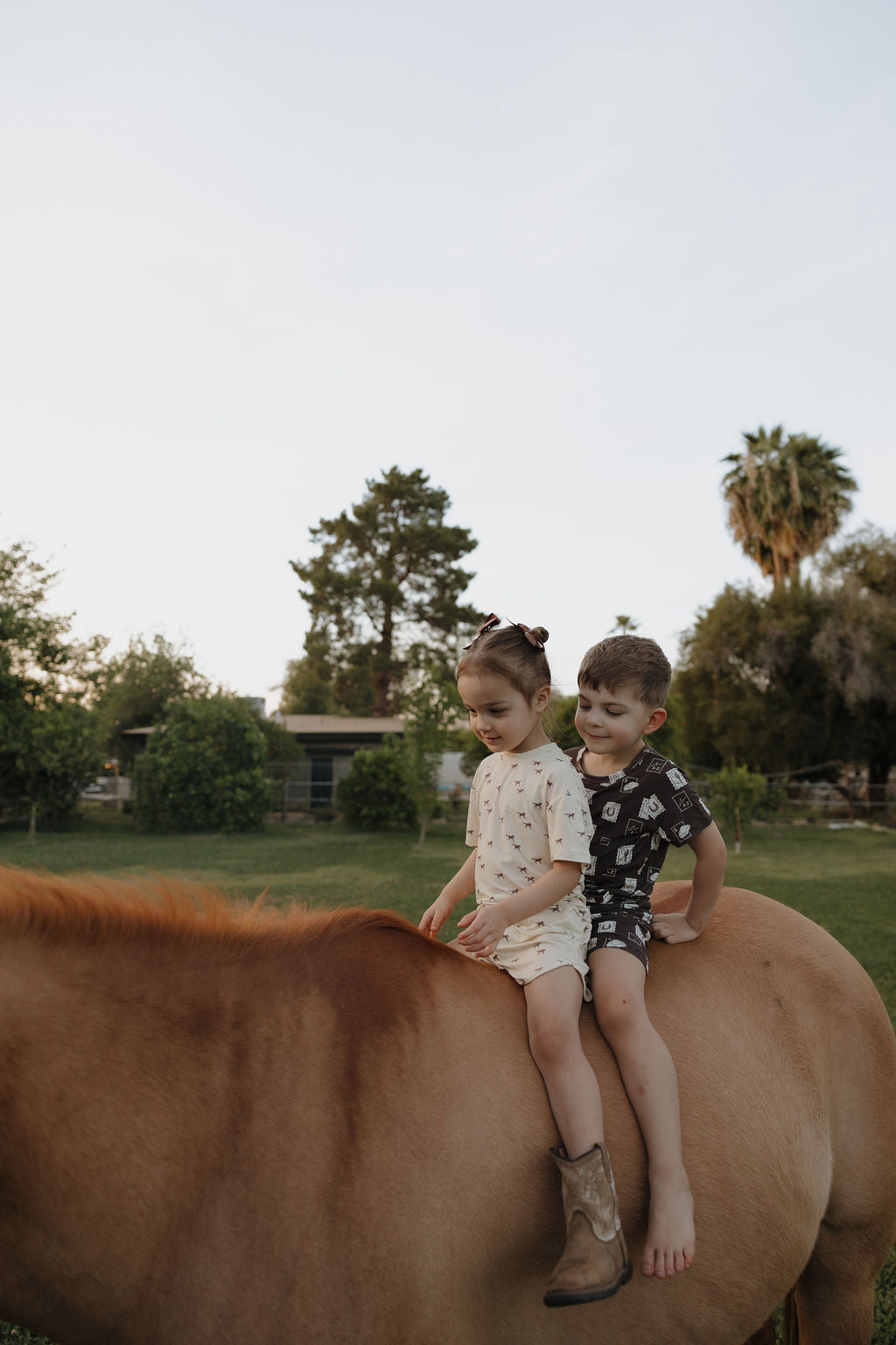 Two children sit on a light brown horse in a grassy yard. One wears the Bamboo Short Set by forever french baby, while the other is dressed in dark clothes. Trees and a house are visible in the background.