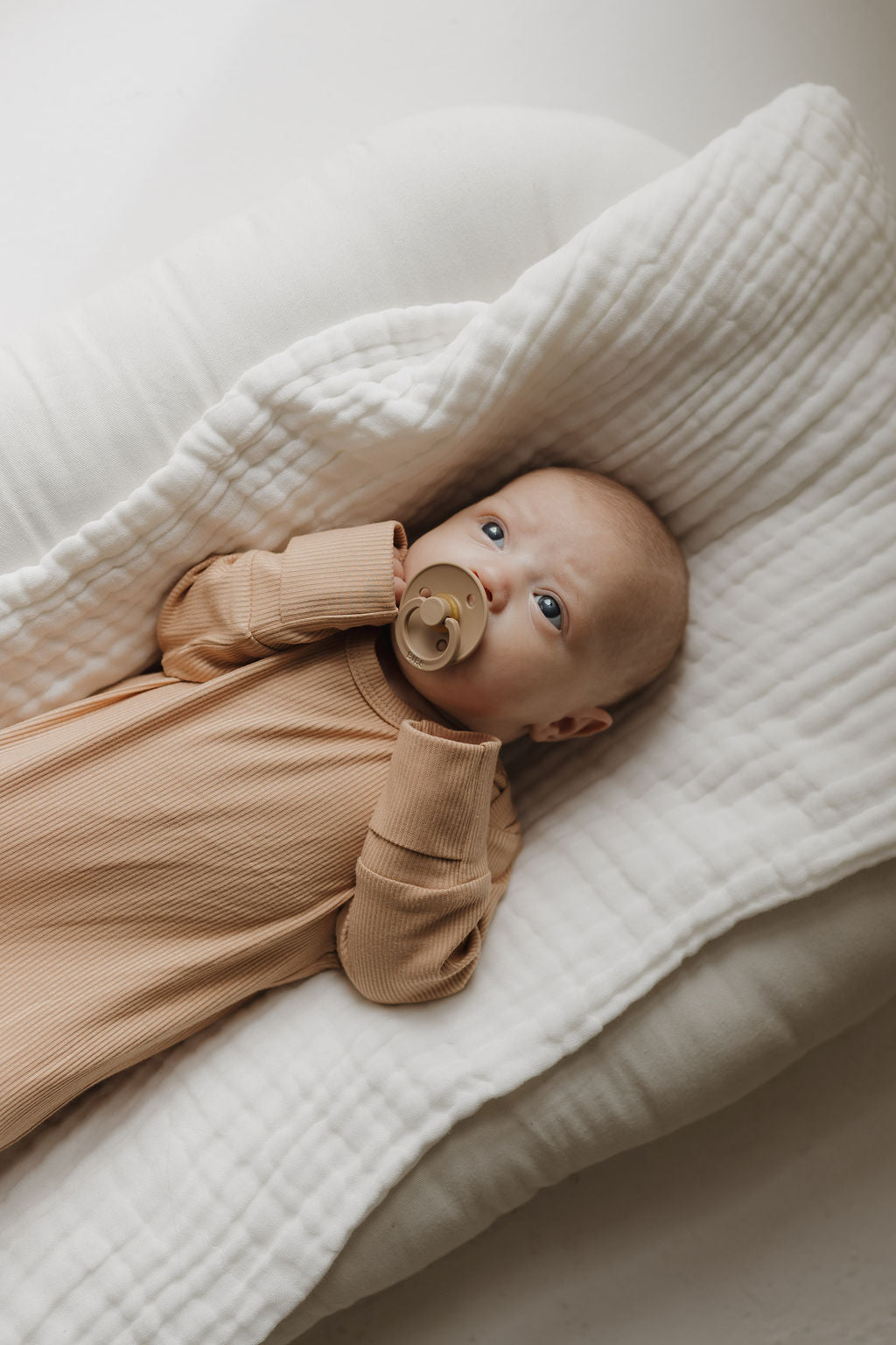 A baby in a forever french baby Bamboo Knotted Gown in Tawny lies on a soft quilted surface with a pacifier, gazing upwards. The softly lit scene creates a calm, serene atmosphere, embodying those newborn essentials moments.