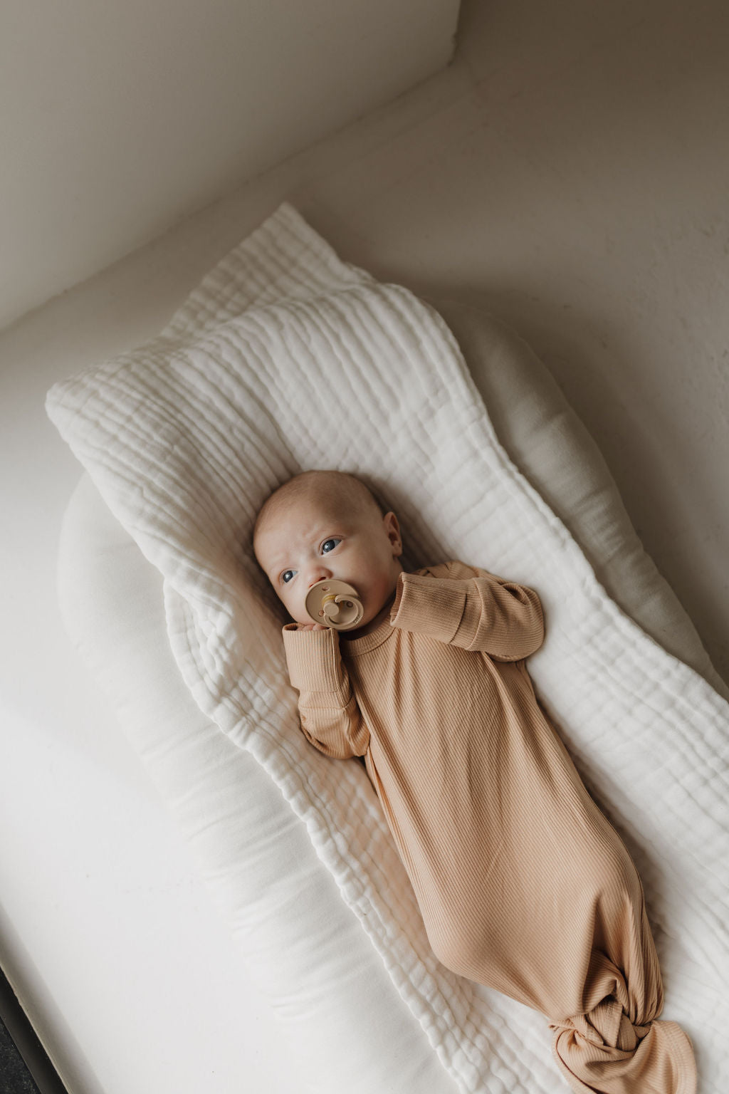 A newborn peacefully lies on a white cushioned pad dressed in the "Bamboo Knotted Gown | Tawny" by forever french baby, holding a pacifier near their mouth. The scene is softly lit, showcasing serene and hypo-allergenic essentials.