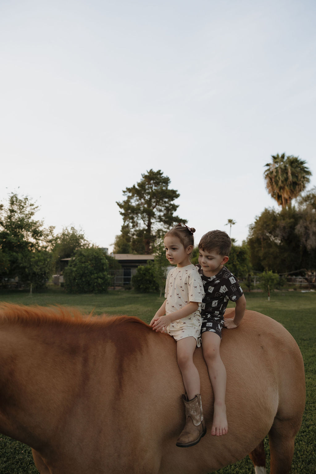 Two young children sit on a brown horse in a grassy yard. The girl in front wears the Girl's Bamboo Short Set | Wild Horses by forever french baby, while the boy behind her is barefoot. Trees and a clear sky are in the background.