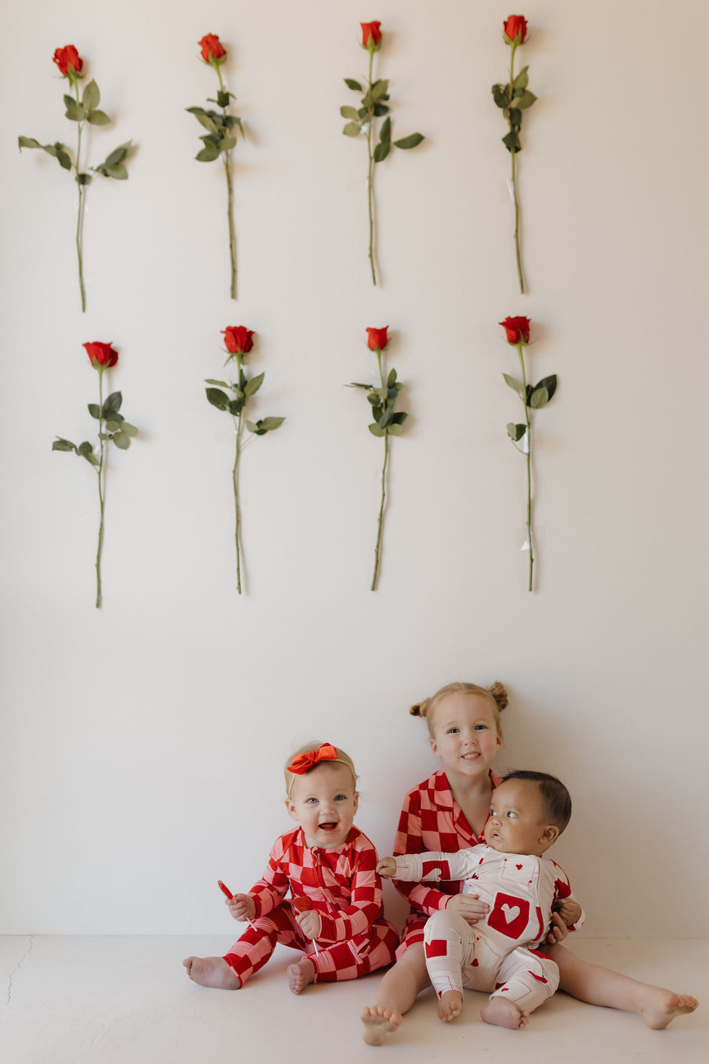 Three children are sitting on the floor wearing matching Bamboo Zip Pajamas in red and white by forever french baby. Behind them, red roses form a grid pattern on the wall. Two children smile at the camera, while the third looks away.