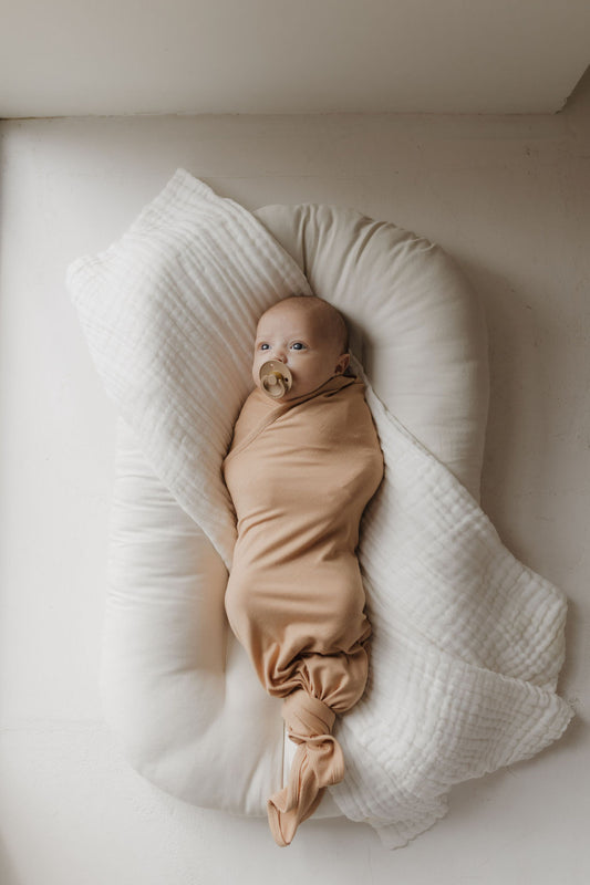 A baby rests softly on a cushion, wrapped snugly in a forever french baby Bamboo Swaddle | Tawny. The serene scene includes the baby looking to the side with a pacifier and a textured white cloth partially covering the cushion.