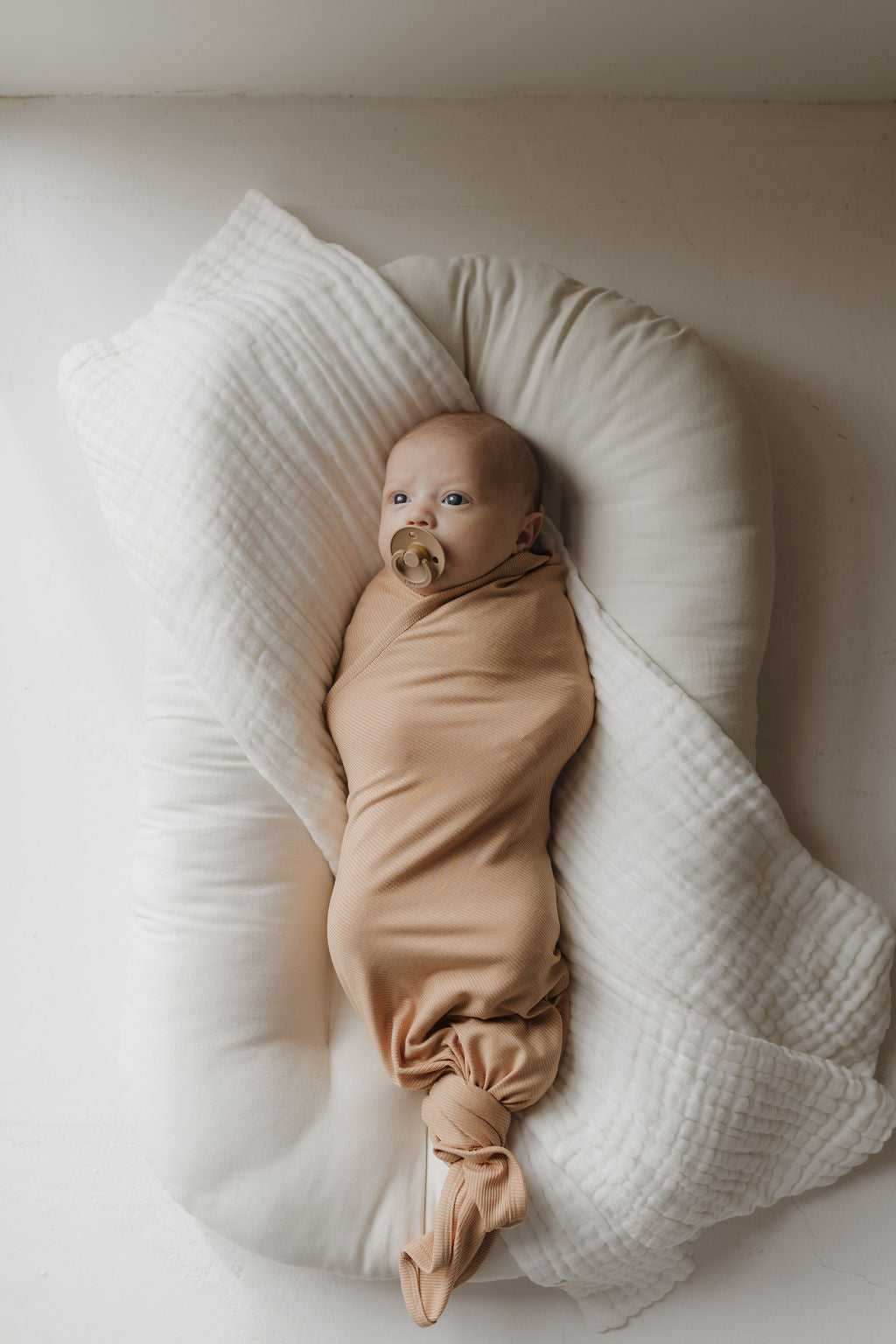 A baby lies awake with a pacifier, wrapped in a Beige Bamboo Swaddle | Tawny by forever french baby, resting on a soft white cushion. The serene scene and gentle lighting highlight the breathable fabric, ensuring comfort for delicate skin.