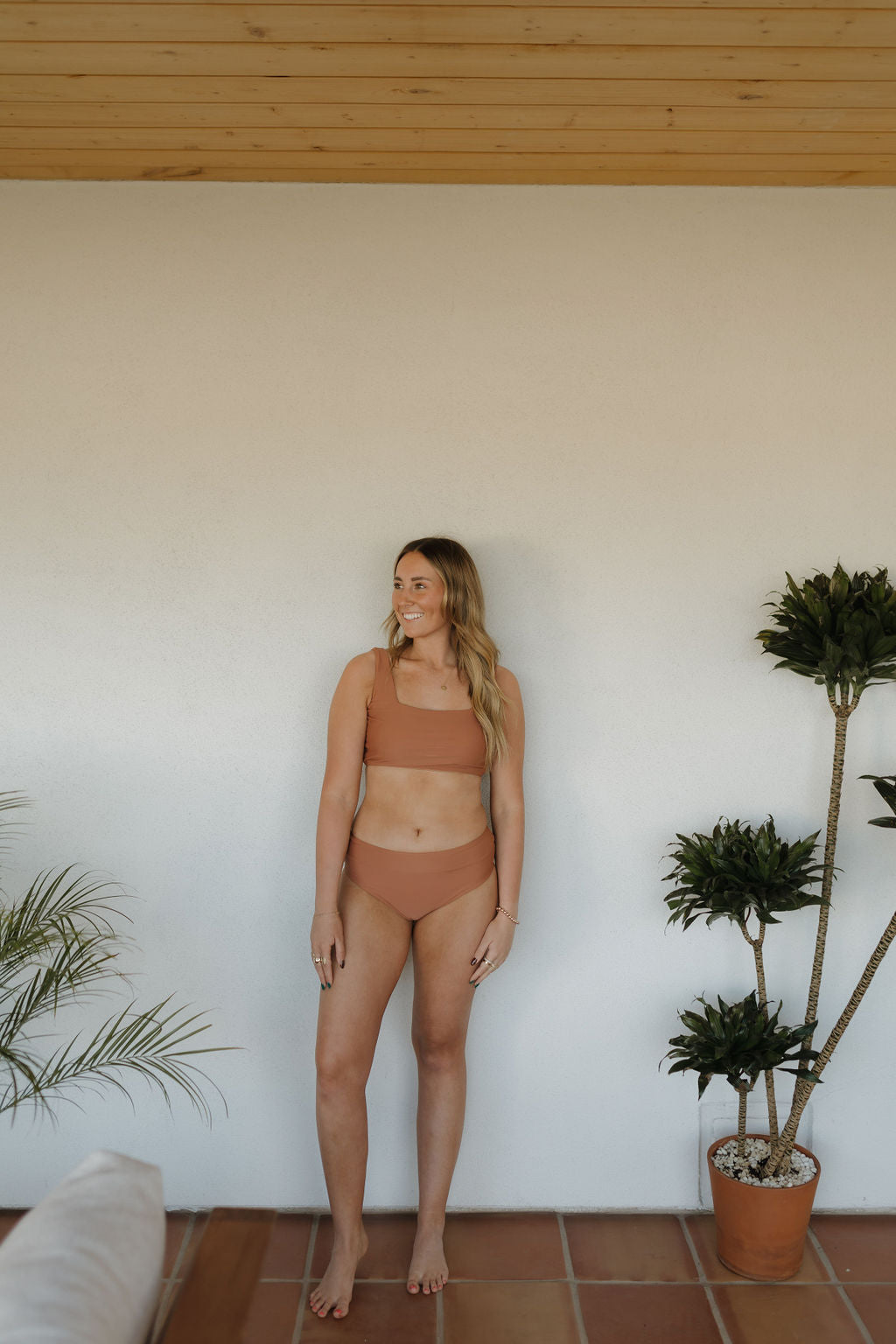 A woman stands barefoot indoors on terracotta tiles, wearing the forever french baby Women's Two Piece Swimsuit from the Terra collection. She smiles with one hand on the wall, surrounded by potted plants that enhance the rustic charm of the wooden ceiling and white wall backdrop.