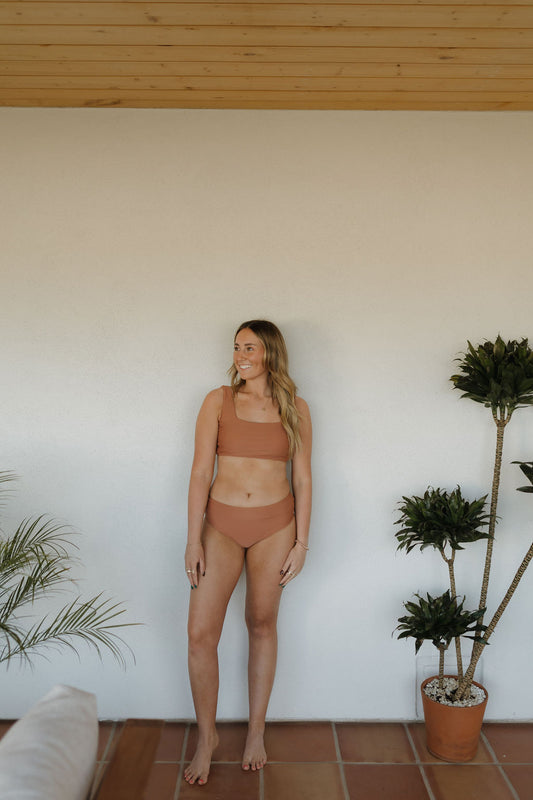 A woman stands barefoot indoors on terracotta tiles, wearing the forever french baby Women's Two Piece Swimsuit from the Terra collection. She smiles with one hand on the wall, surrounded by potted plants that enhance the rustic charm of the wooden ceiling and white wall backdrop.