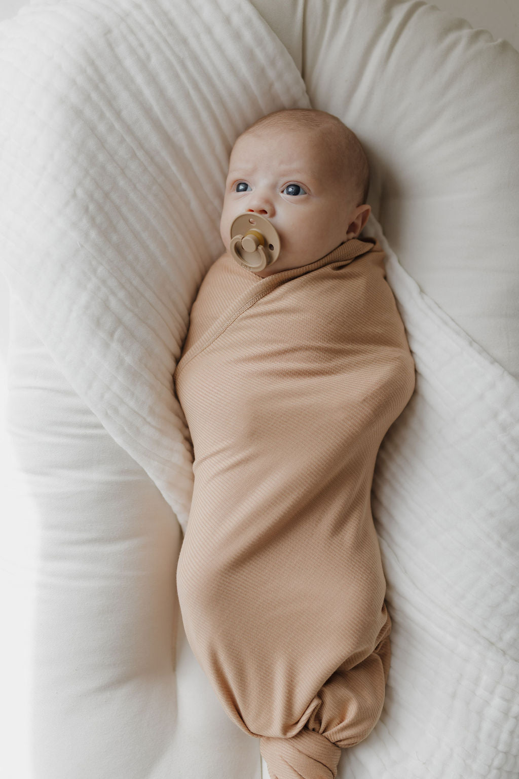 A baby lies swaddled in the forever french baby Bamboo Swaddle | Tawny, resting on a soft, white cushion. The breathable fabric envelops the little one, who has a pacifier and gazes upward with wide eyes.