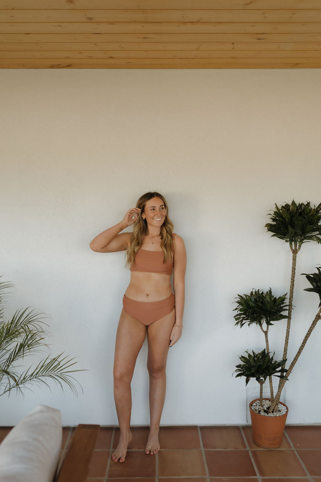 A woman in a rust-colored Women's Two Piece Swimsuit from forever french baby’s Terra collection stands indoors on terracotta tiles, smiling and glancing to the side, surrounded by potted plants with a wooden ceiling above and a white wall behind her.