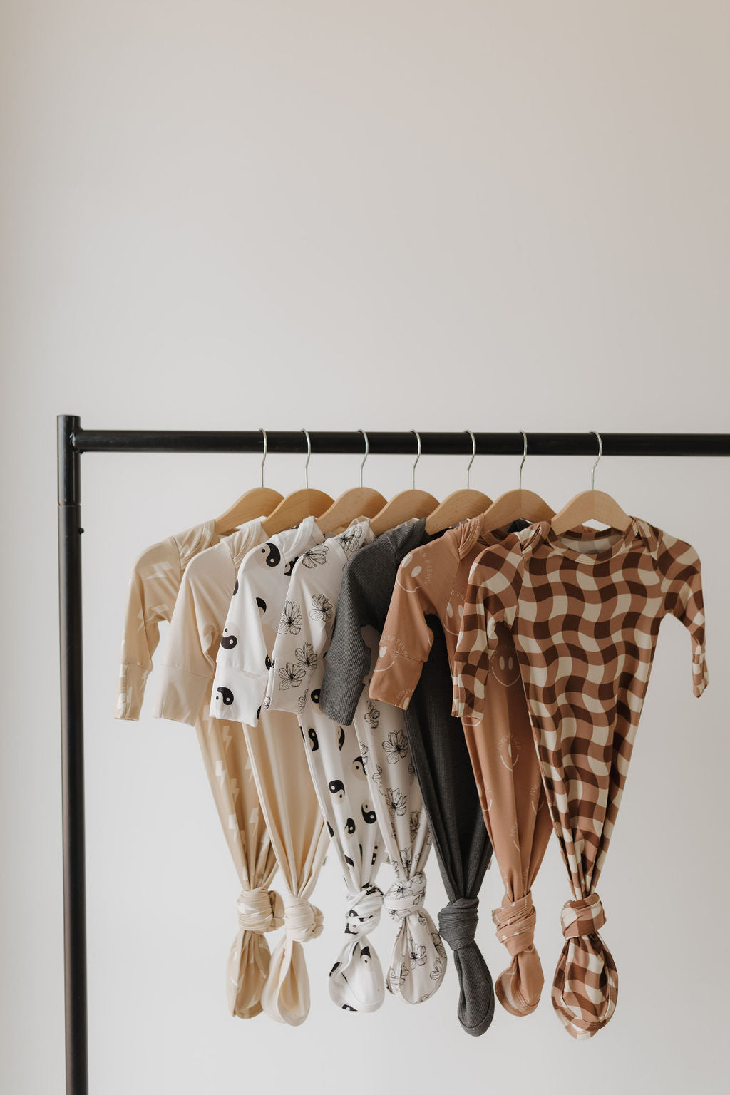 Seven "Bamboo Knotted Gown | Black & White Yin Yang" gowns by forever french baby, feature knotted bottoms for easy adjustment. These neutral-toned, hypo-allergenic essentials hang on wooden hangers from a black rack and include prints like polka dots, checkered, and abstract designs.