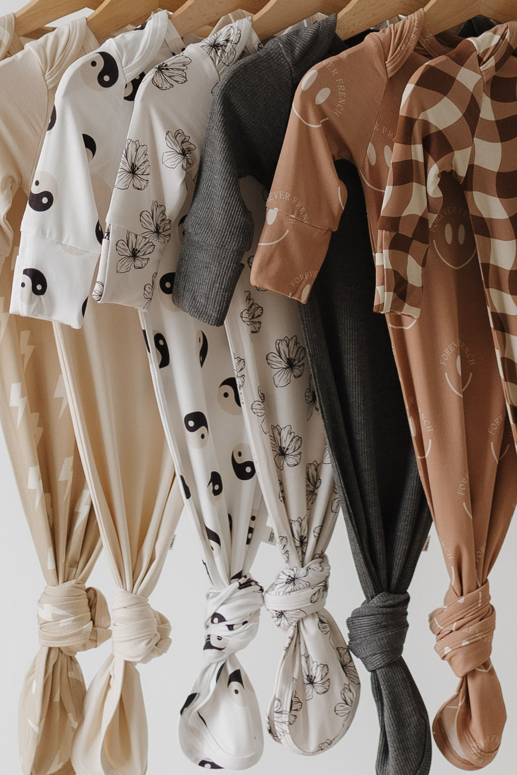 A row of forever french baby's Bamboo Knotted Gowns hangs on a wooden rod, each featuring hypo-allergenic essentials with black and white yin-yang patterns in neutral tones of beige, cream, gray, and brown. Stylishly knotted at the bottom, these gowns are newborn must-haves.