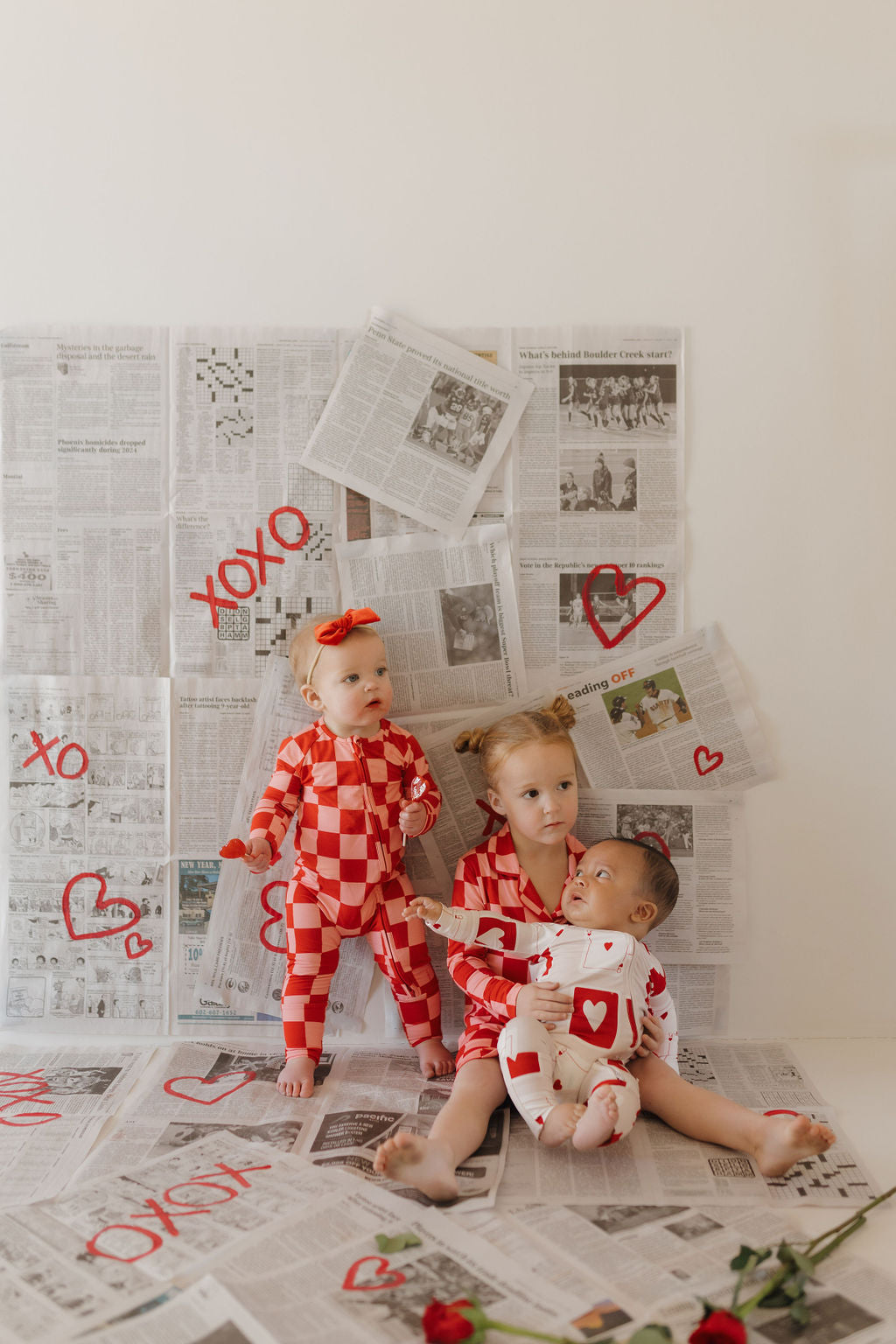 Three children in forever french baby's Bamboo Two Piece Pajamas | XOXO sit on a newspaper-covered floor. Painted red hearts and "XOXO" adorn the papers, while a red rose adds warmth to their cozy scene.