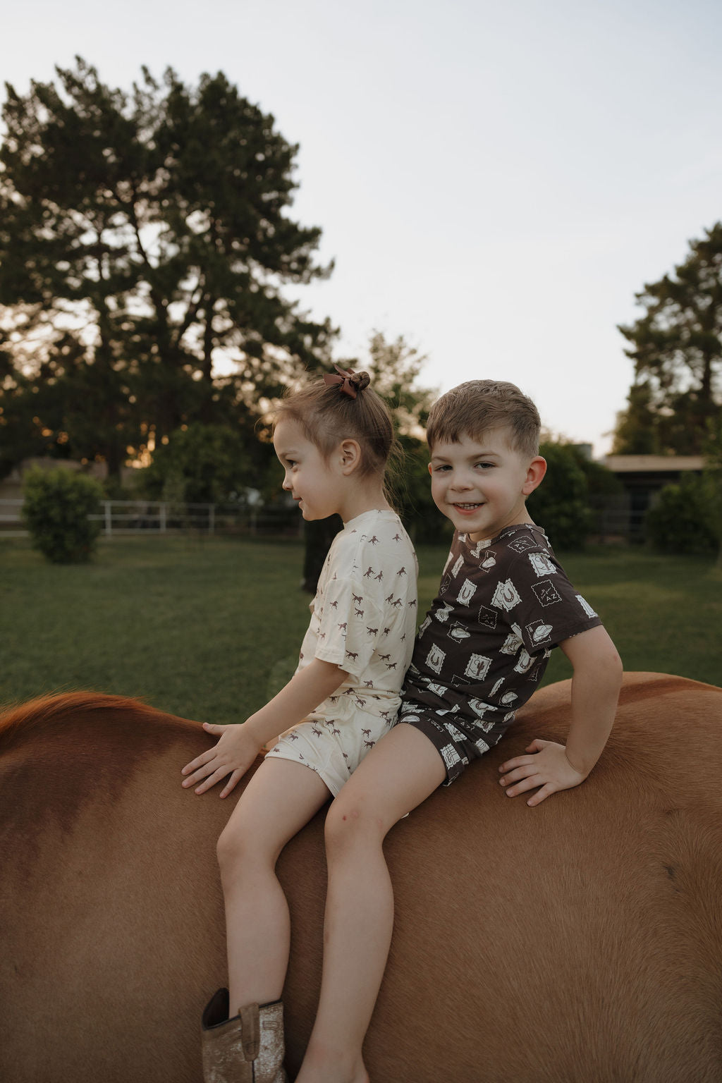 Two children sit on a brown horse outdoors. The boy, wearing forever french baby’s Bamboo Short Set | With Love from AZ, smiles at the camera while trees and grass fill the background. The girl sits beside him facing forward.