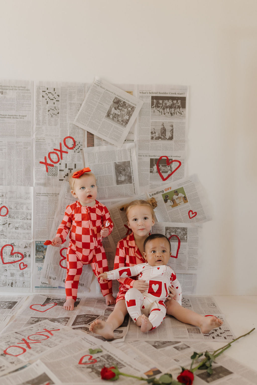 Three children wearing forever french baby's Bamboo Two Piece Pajamas | XOXO sit on a newspaper-covered floor. Red hearts and "XOXO" are painted on the papers, with two red roses beside them. The wall is also covered in newspapers.