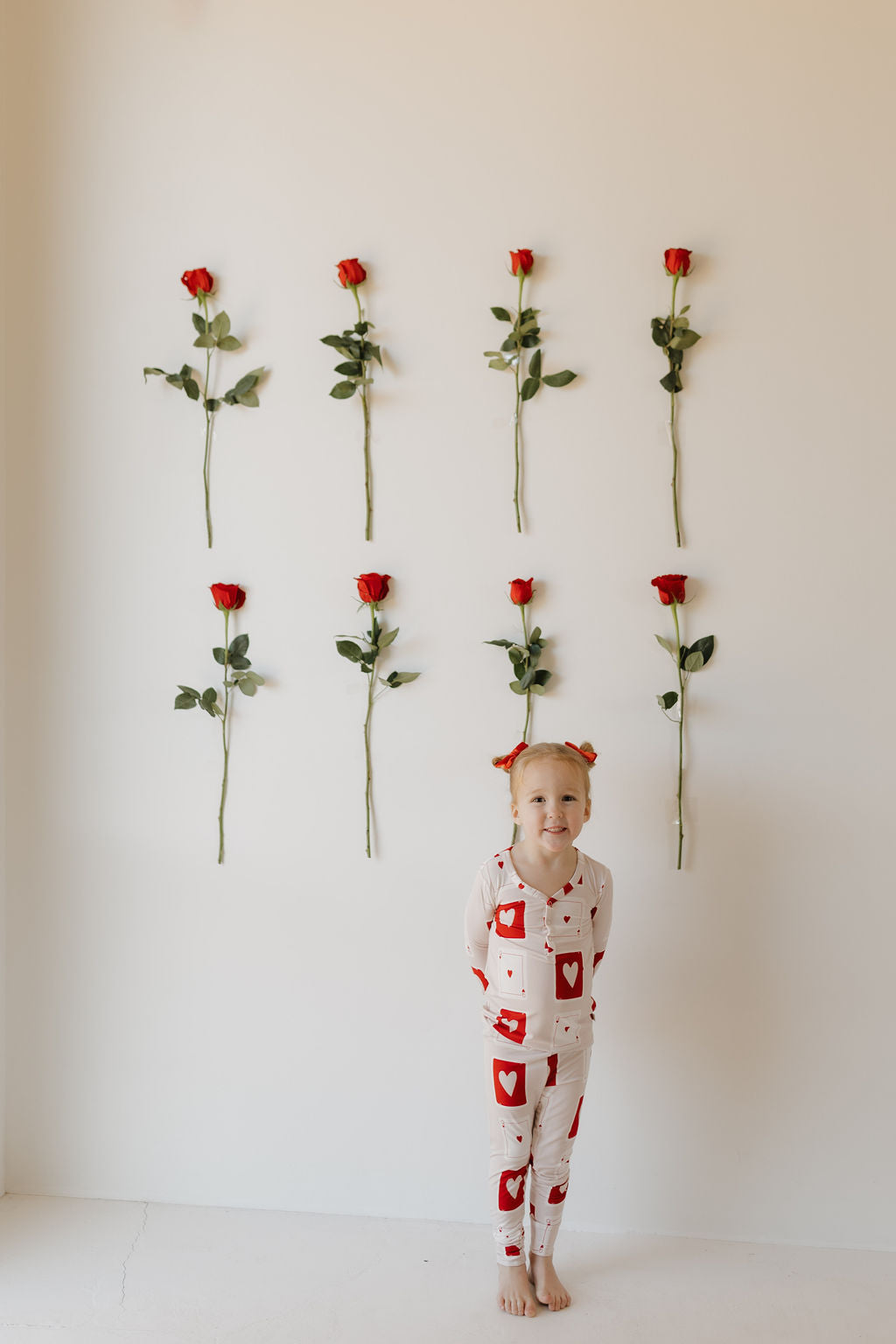 A child smiles barefoot in forever french baby's "Love Day" Bamboo Two Piece Pajamas, featuring heart patterns. Against a white backdrop with six vertically arranged red roses, the minimalist setting accentuates the breathable fabric ideal for tender skin.