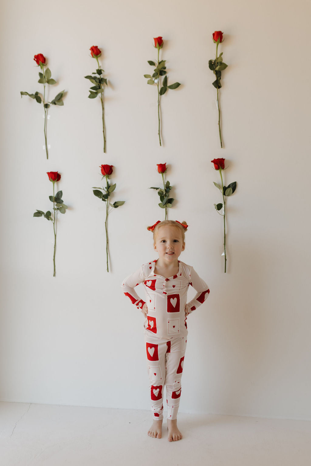 A child in forever french baby's Love Day bamboo pajamas stands confidently, hands on hips, smiling in front of a white wall adorned with nine vertical red roses.