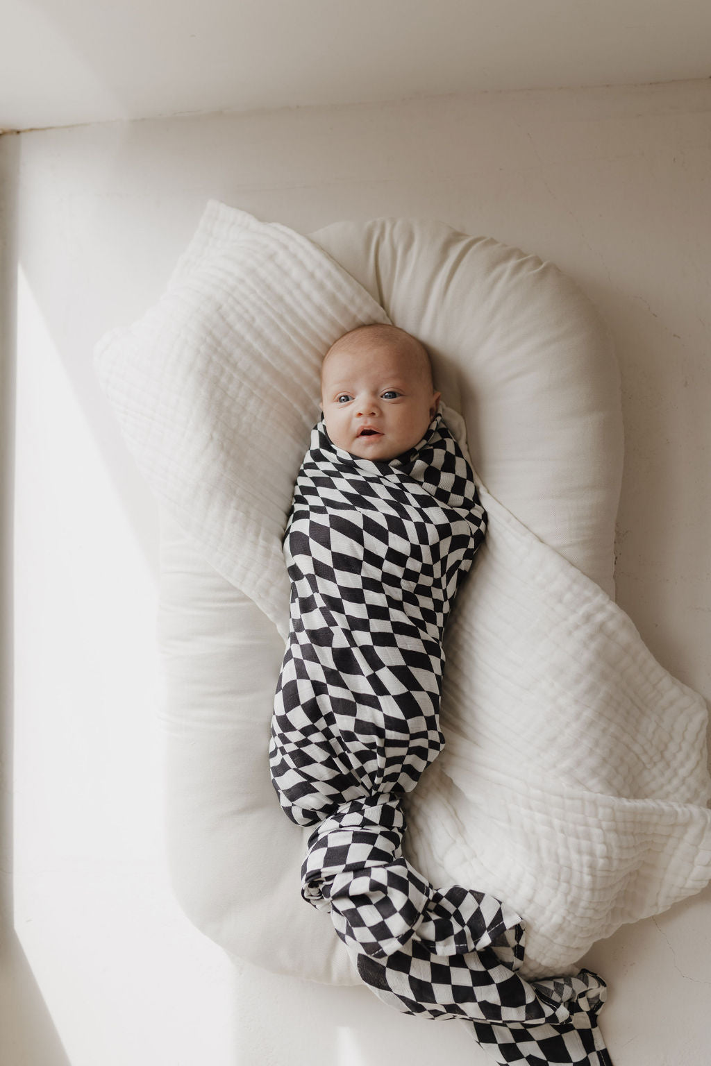 A baby rests on a white cushion, wrapped in an ultra-soft Muslin Swaddle from forever french baby, featuring a black and white wavy checkerboard design. Sunlight streams in from the left, casting gentle shadows as the baby looks up with wide-open eyes, embraced by 100% Muslin Cotton coziness.