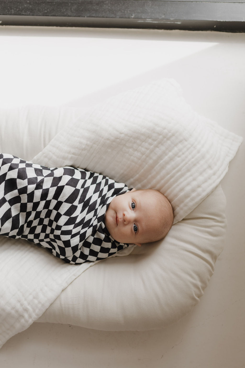 A baby rests on a soft, white cushion, wrapped in forever french baby's ultra-soft Muslin Swaddle in Black & White Wavy Checkerboard design. The child gazes upwards with big eyes, illuminated by gentle natural light from a nearby window.