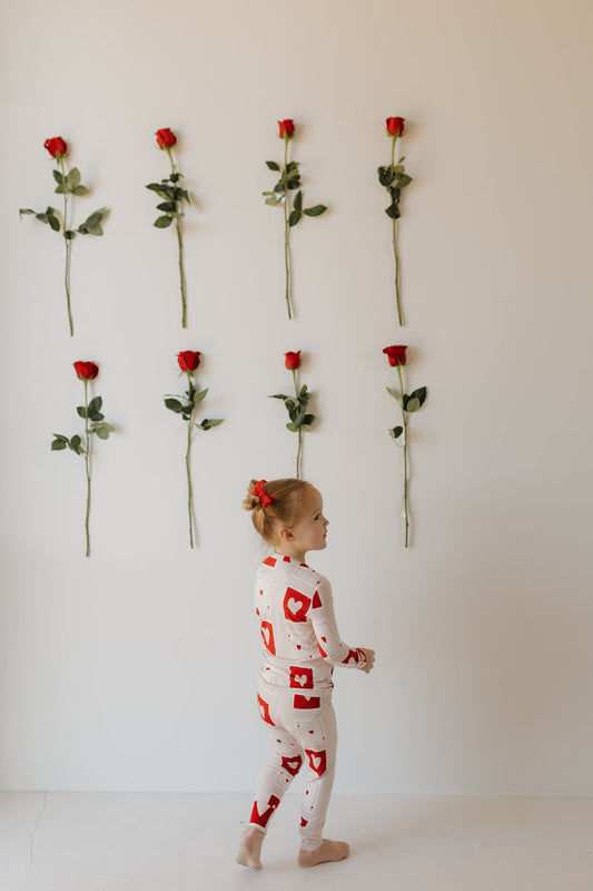 A child wears the "Bamboo Two Piece Pajamas | Love Day" by forever french baby, patterned with hearts. They face a wall with eight red roses in two rows, and their hair is tied with a red bow, complementing the room's soft neutrals. The breathable fabric ensures comfort as they stand quietly.