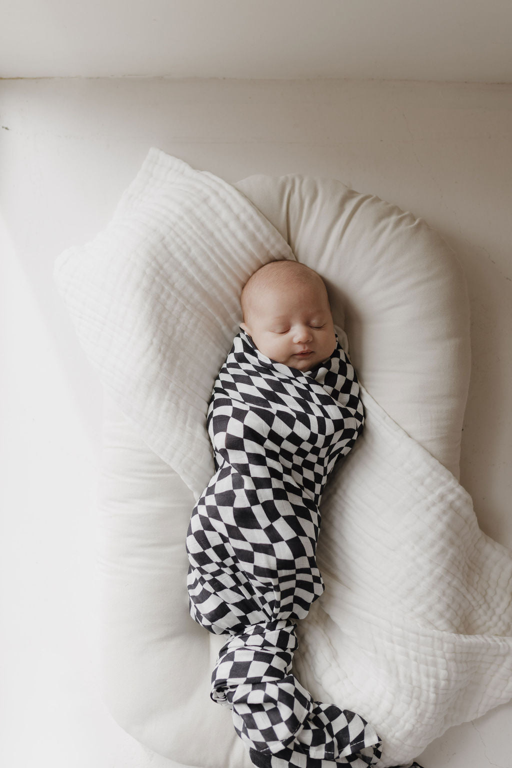 A baby sleeps peacefully, wrapped in a forever french baby Muslin Swaddle in Black & White Wavy Checkerboard. The 100% cotton blanket lies on a cushioned white surface, creating a serene and airy atmosphere.